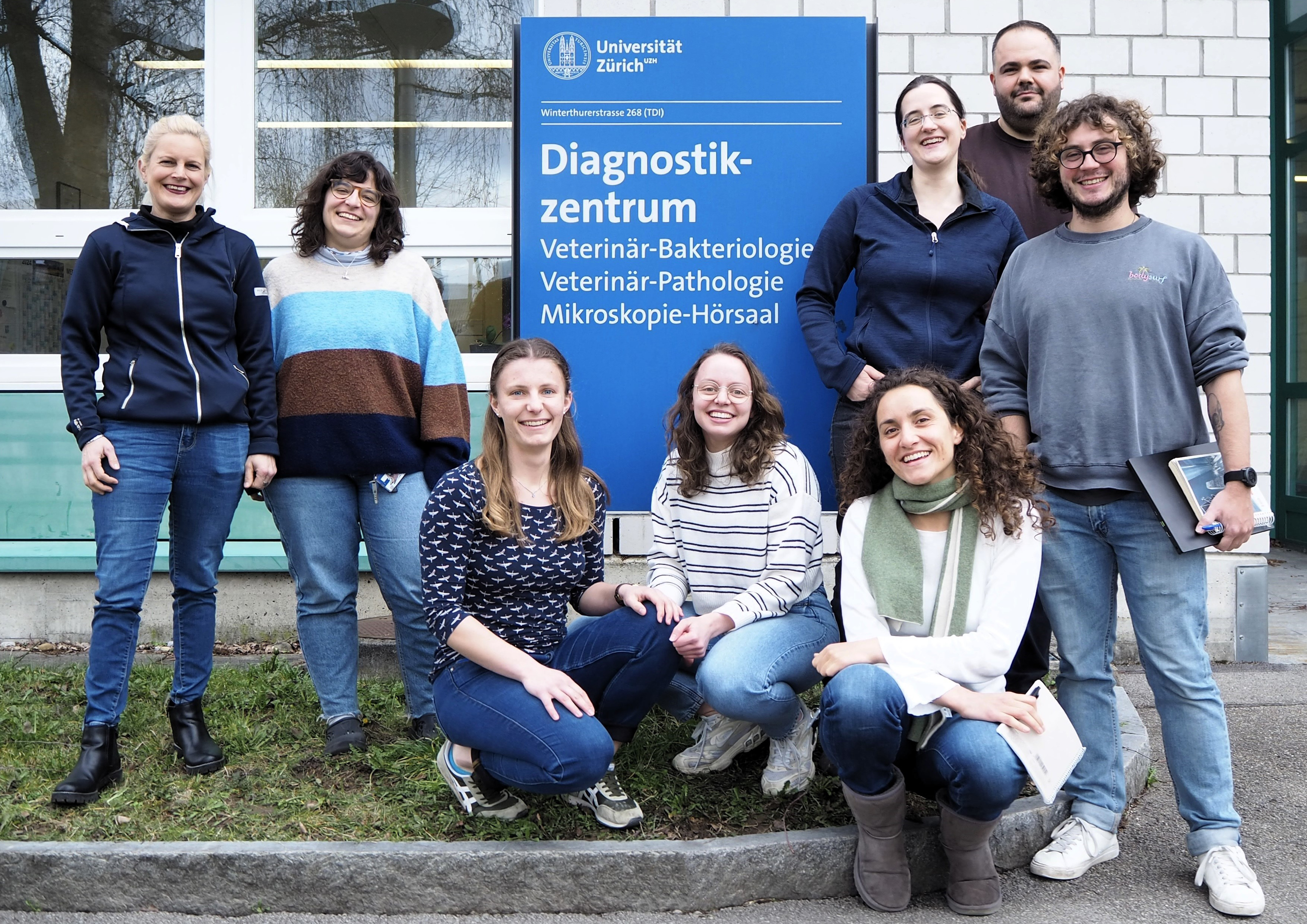 A group photo of the ChlamHealth team, from left to right: Nicole Borel, Magdalena de Arriba Sanchez de la Campa, Daphne Zubler, Nadja Fässler, Giuliana Malingamba, Hanna Marti, Efe Altuntas, and Enrique Rayo, standing in front of the “Diagnostikzentrum” sign at Vetsuisse UZH.